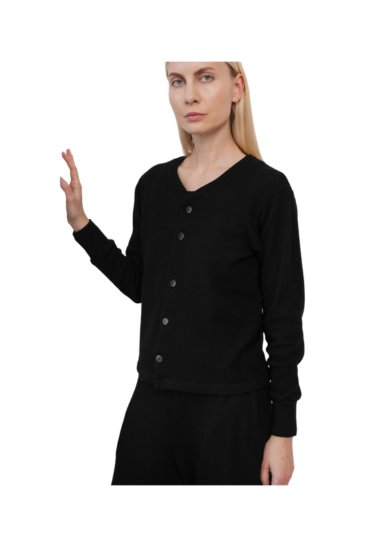 A woman with straight blonde hair models the Wol Hide Wool Liner in Onyx—a minimalist black cardigan—paired with black pants against a white background, showcasing a modern, sustainable style as she raises her left hand.