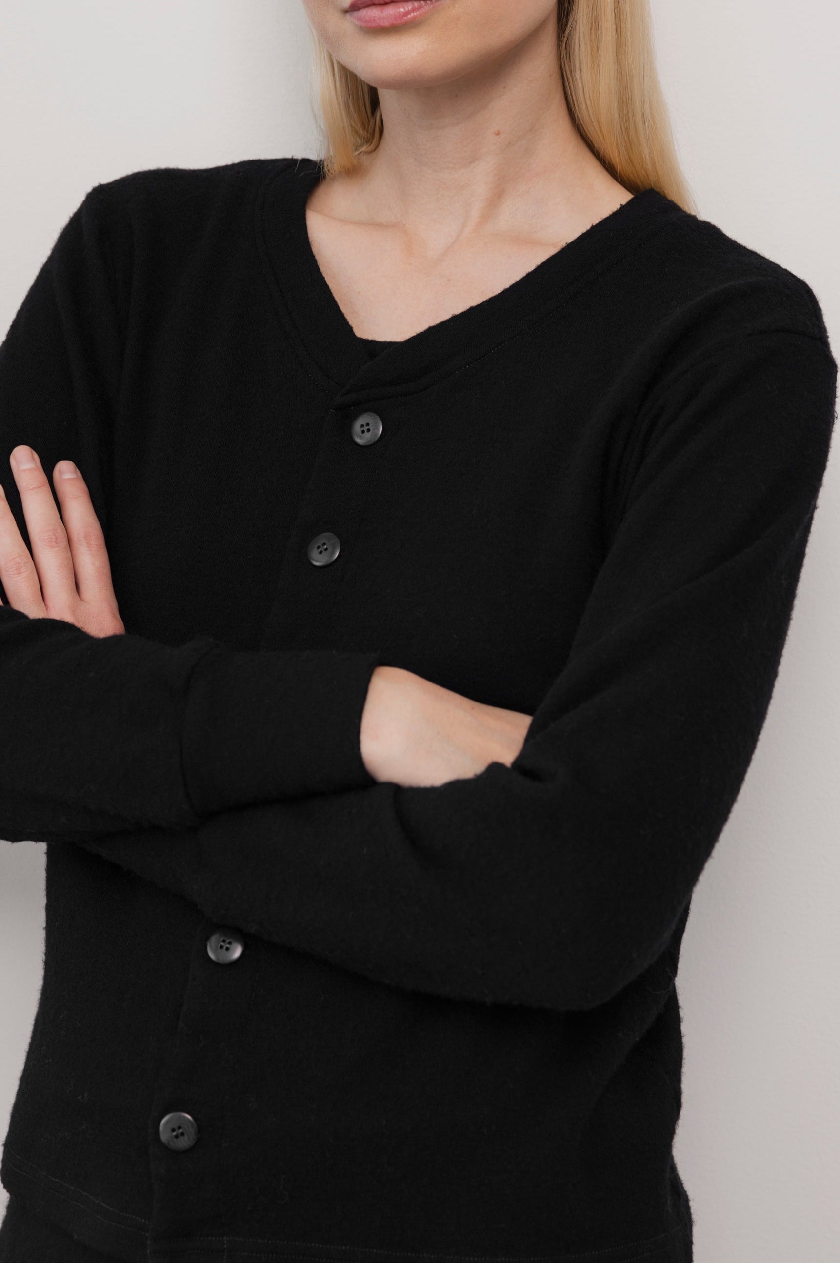 A person with long blonde hair stands with arms crossed, wearing the Wol Hide Wool Liner : Onyx cardigan with Corozo buttons. The cropped image shows just their torso and lower face against a plain light background.