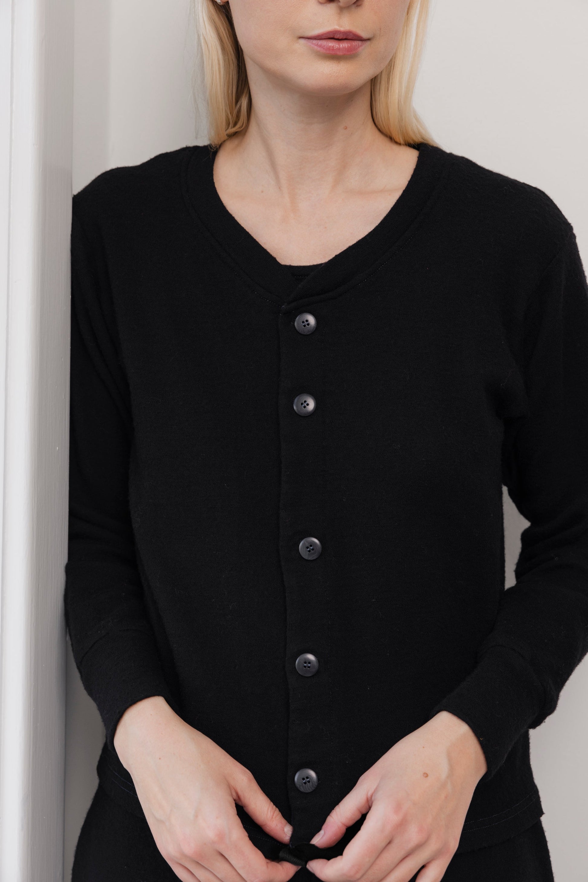 A woman with light skin and blonde hair stands against a white wall wearing the Wol Hide Wool Liner in Onyx, a black slim cardigan with Corozo buttons. Her hands are clasped near the bottom and her face is partially visible, eyes out of frame.