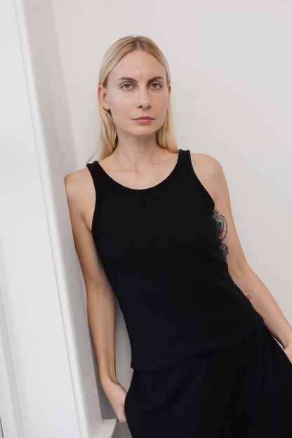 A woman with long blonde hair stands indoors against a white wall, wearing the Wol Hide Wool Tank in Onyx and black pants. She looks directly at the camera with a neutral expression; a side tattoo is partially visible.