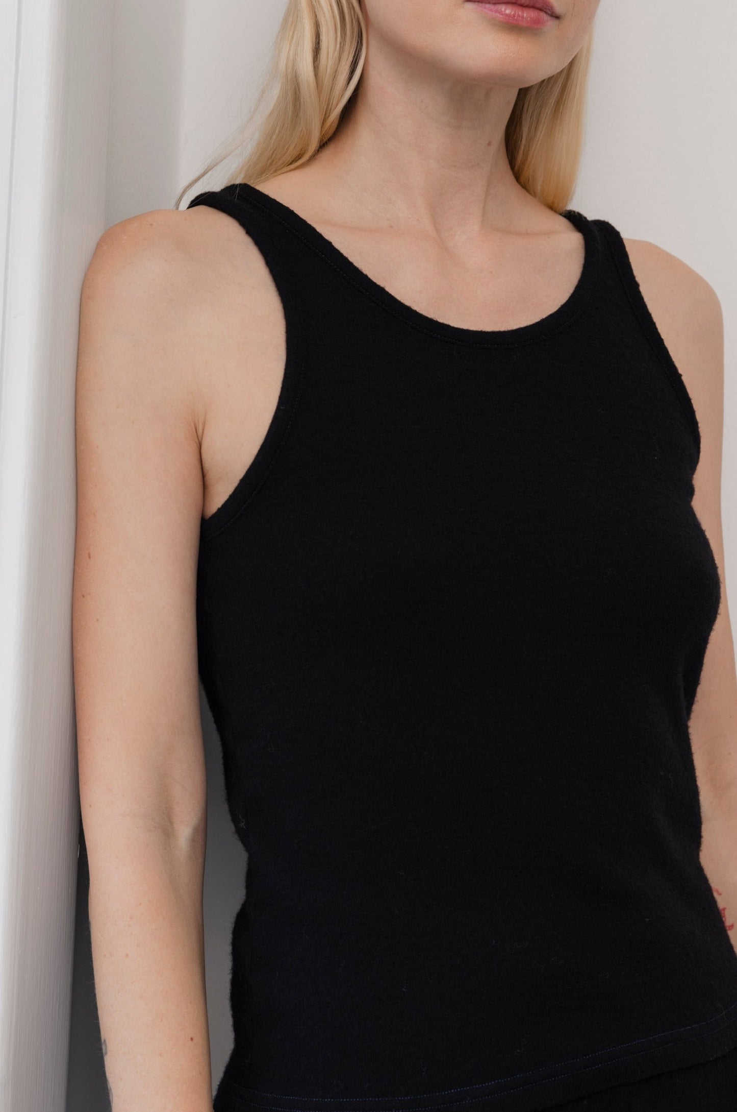 A woman with long blonde hair models the Wol Hide Wool Tank: Onyx, a sleeveless black layering top, standing against a white wall; only her lower half and torso appear in the frame.
