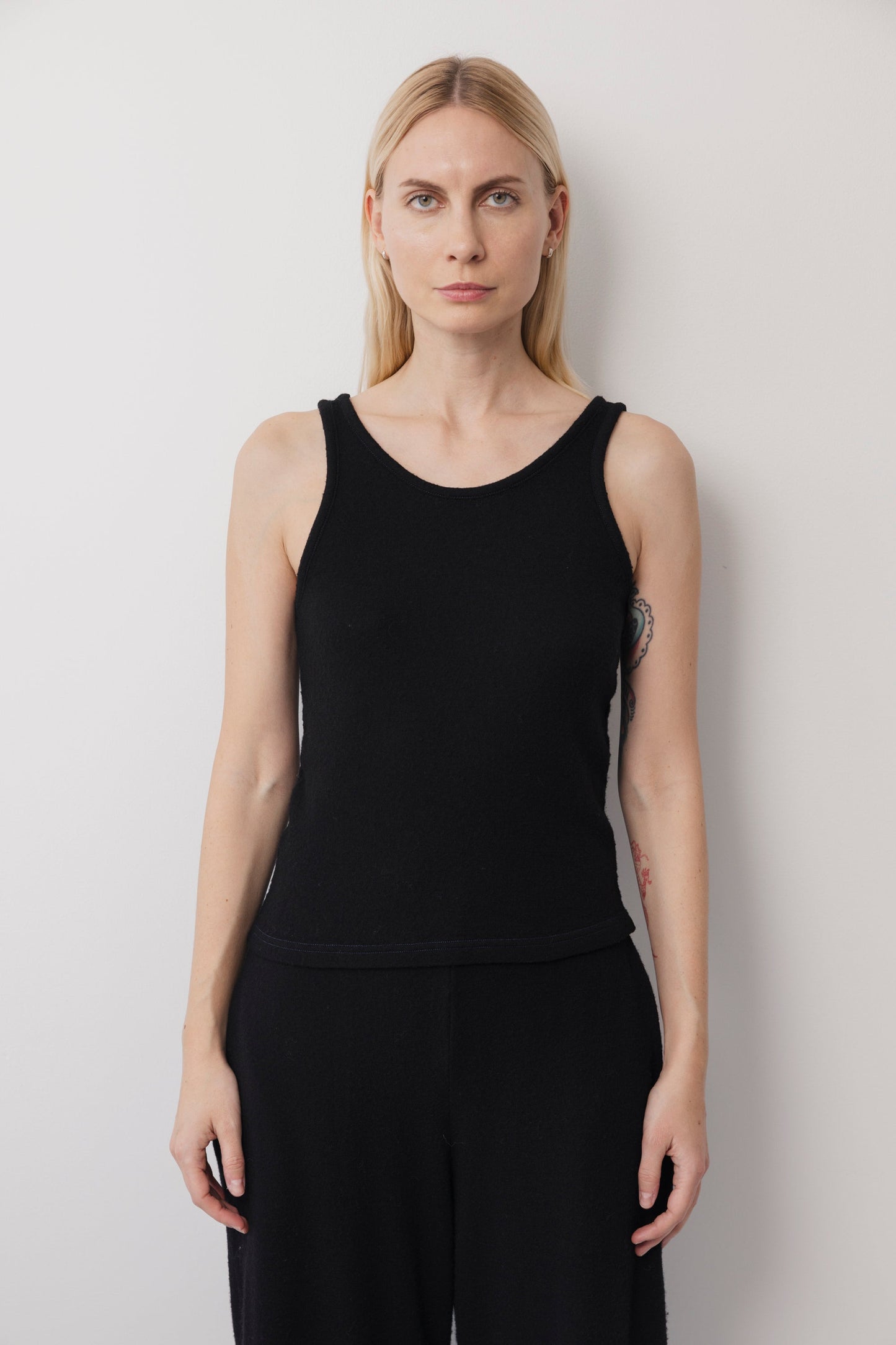 A woman with straight blonde hair wears the Wol Hide Wool Tank: Onyx with black pants, standing against a white background and facing the camera with a neutral expression.