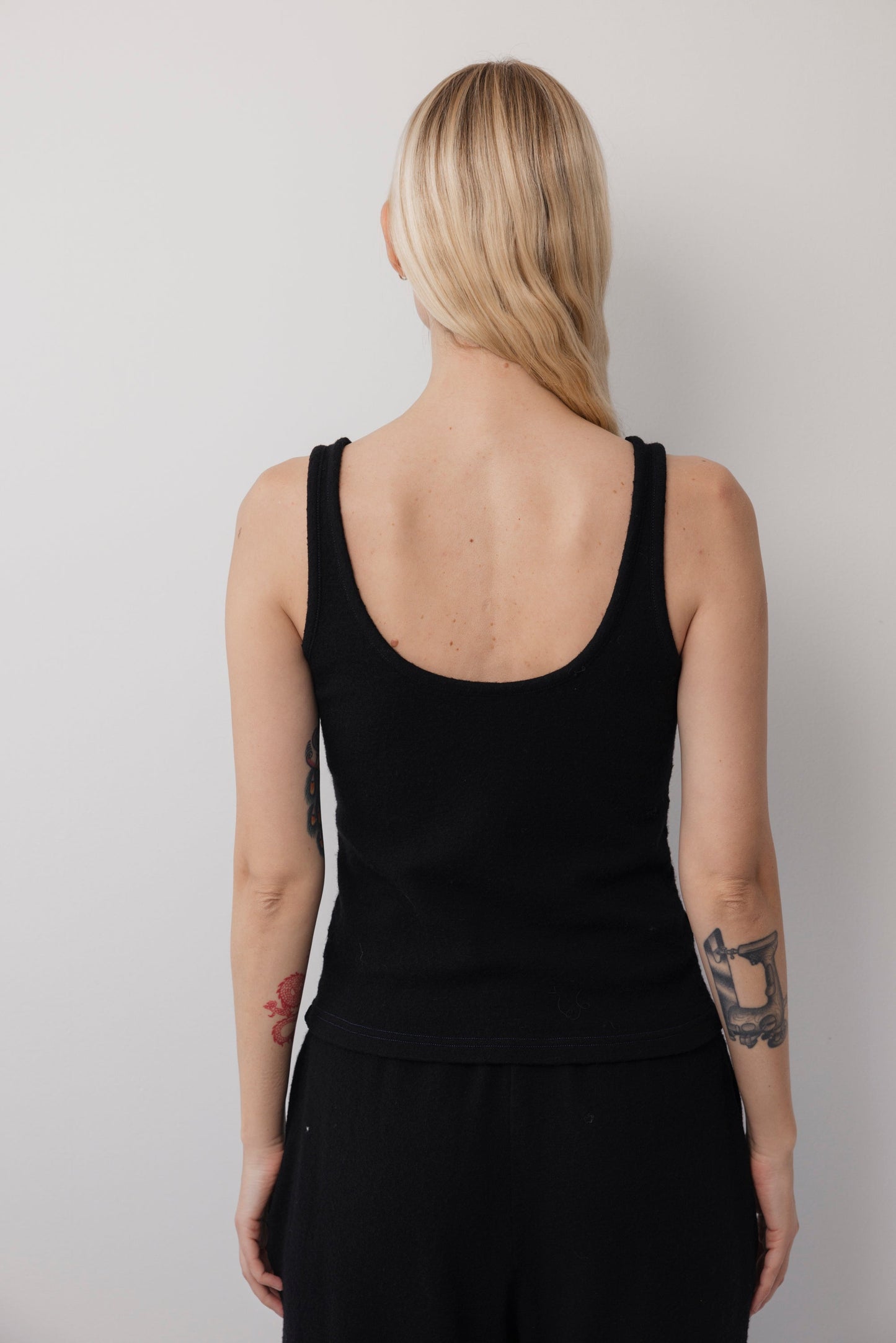 A woman with long blonde hair and visible arm tattoos stands against a light wall, seen from behind wearing the Wol Hide Wool Tank: Onyx and black pants. The tank features a low scoop back for effortless layering.