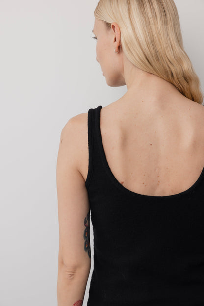 A woman with long blonde hair faces away from the camera, showing her back and shoulder in the Wol Hide Wool Tank: Onyx, a sleeveless black layering piece. A tattoo is visible on her upper arm against a light, plain background.