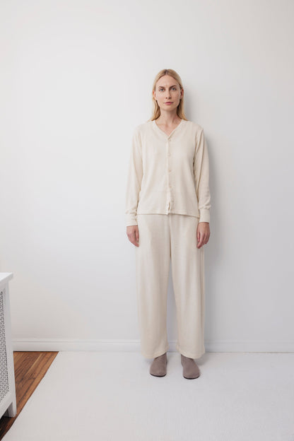 A blonde woman stands against a white wall, wearing Wol Hide's Wool Liner : Natural cream cardigan with Corozo buttons and matching wide-leg pants, paired with gray slippers on a white carpet over a small wood floor section.