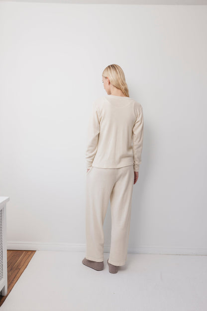 A person with long blonde hair stands with their back to a white wall in a minimal, bright room, wearing the Wol Hide Wool Liner : Natural—a slim, lightweight wool cardigan with Corozo buttons. Their hands rest at their sides.