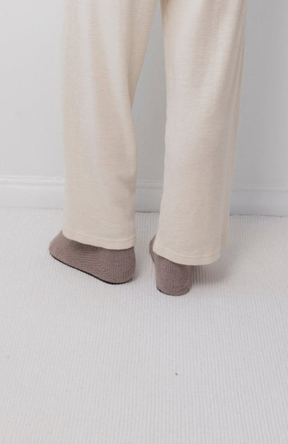 A person wearing Wol Hide's Wool Wide Leg: Natural pants and brown socks stands on a light carpet near a white wall, with only their lower legs and feet visible.