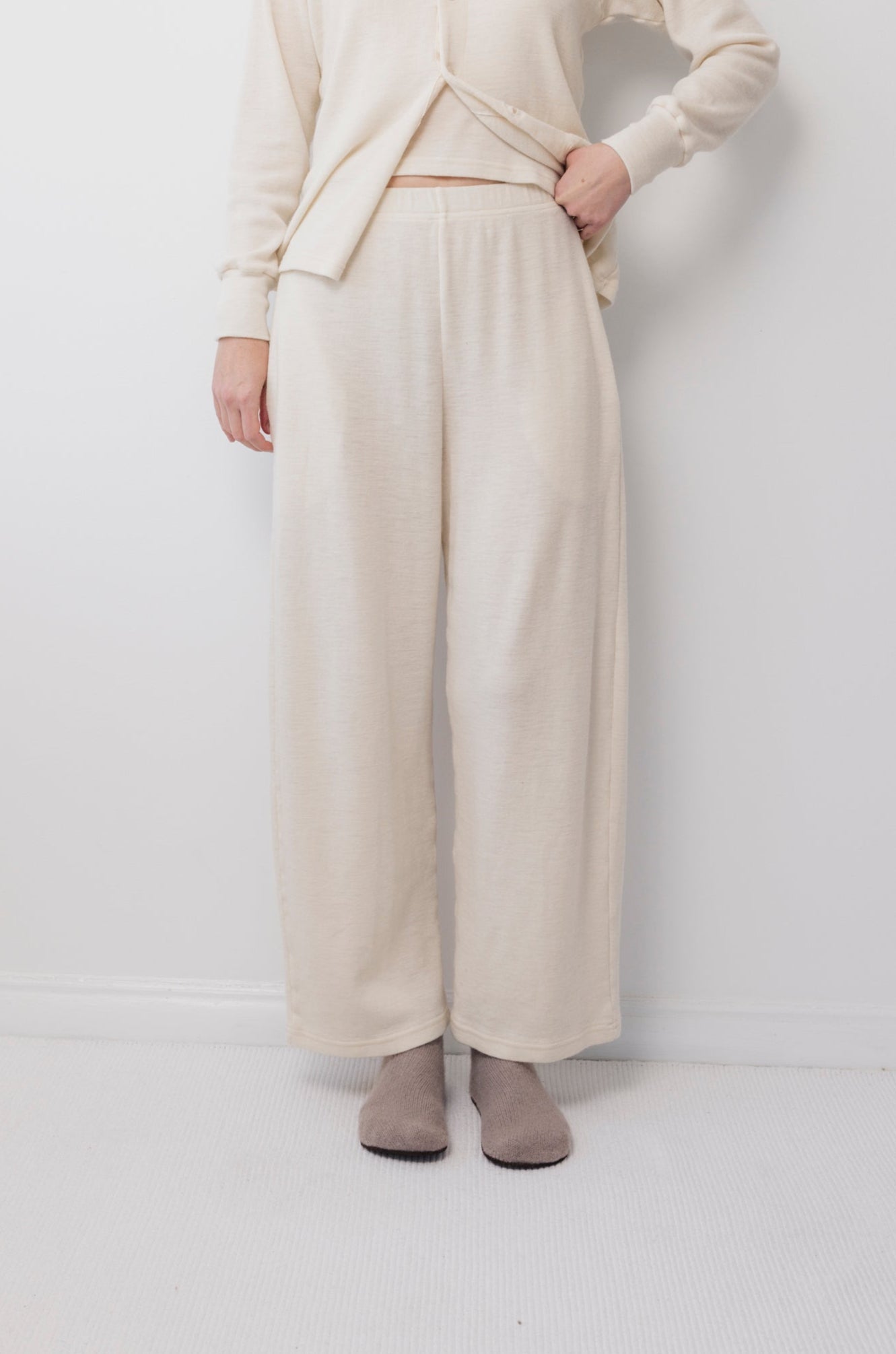Someone wearing Wol Hide's Wool Wide Leg: Natural pants and a matching top stands against a white wall and carpet, holding up the shirt slightly to reveal the elastic waist. Light gray socks complete the look.