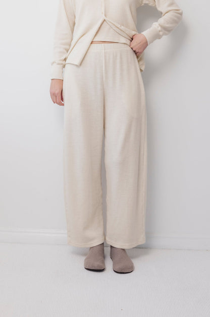 Someone wearing Wol Hide's Wool Wide Leg: Natural pants and a matching top stands against a white wall and carpet, holding up the shirt slightly to reveal the elastic waist. Light gray socks complete the look.