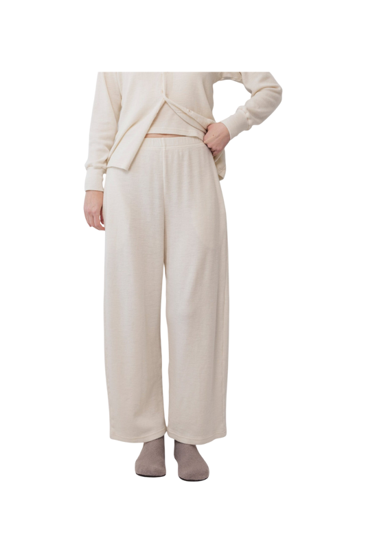 A person wears Wol Hide’s Wool Wide Leg: Natural lounge pants with a matching top and gray slippers, standing against a white background. Their hand is on their hip, and their face is out of frame.