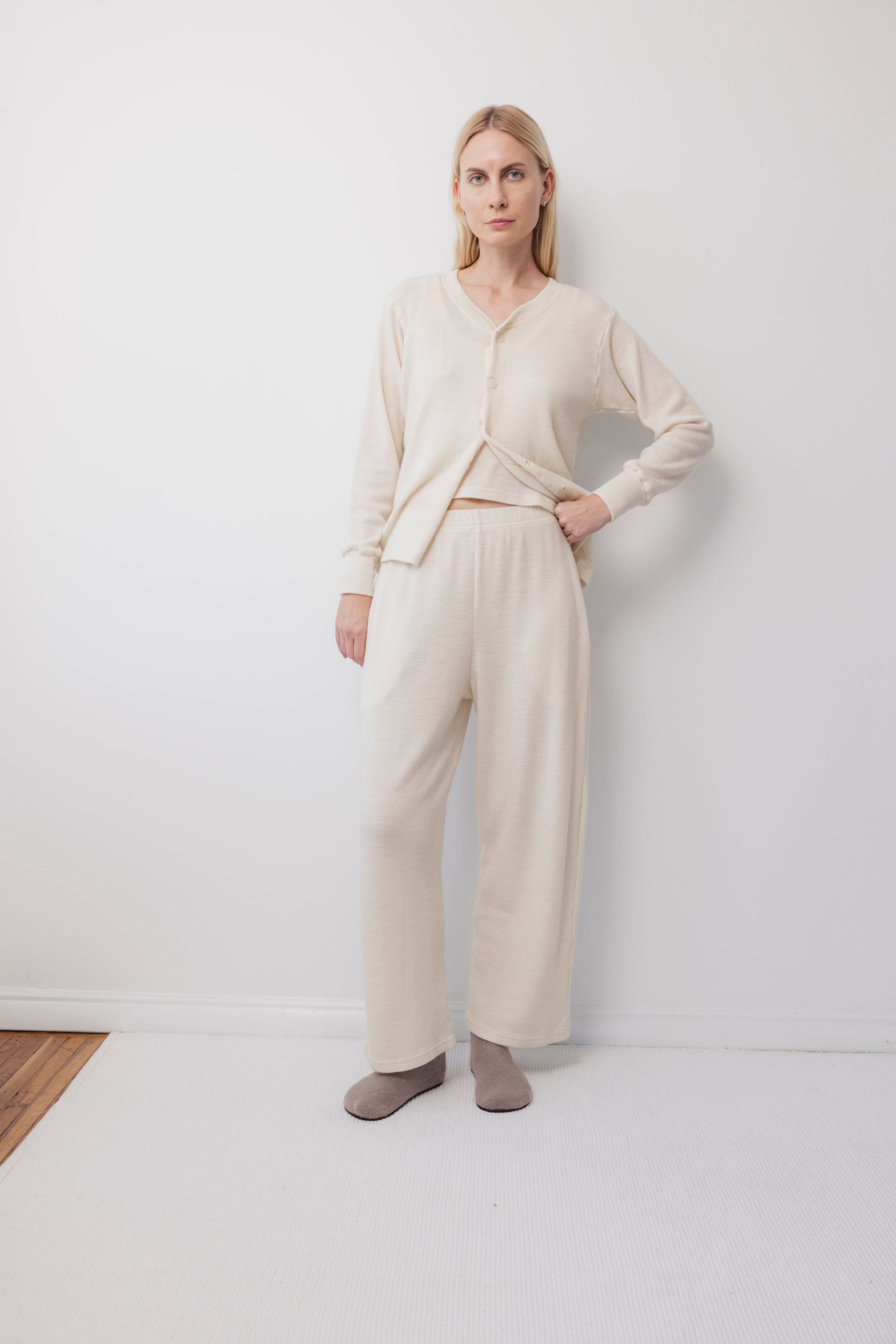 A person stands indoors against a white wall, wearing the Wol Hide Wool Liner : Natural slim cardigan with Corozo buttons and matching wide-leg pants, paired with taupe slippers. One hand is in their pocket and they have a neutral expression.