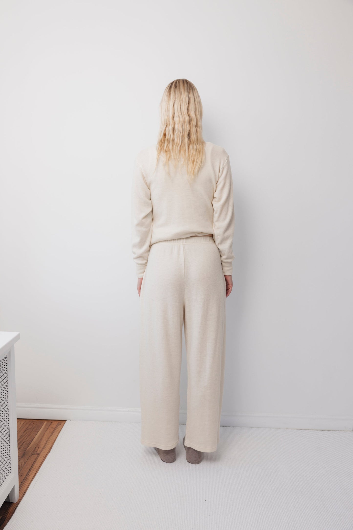A person with long blonde hair stands facing a white wall, wearing Wol Hide's Wool Wide Leg: Natural pants and a matching cream-colored long-sleeve top, on a white carpeted floor.