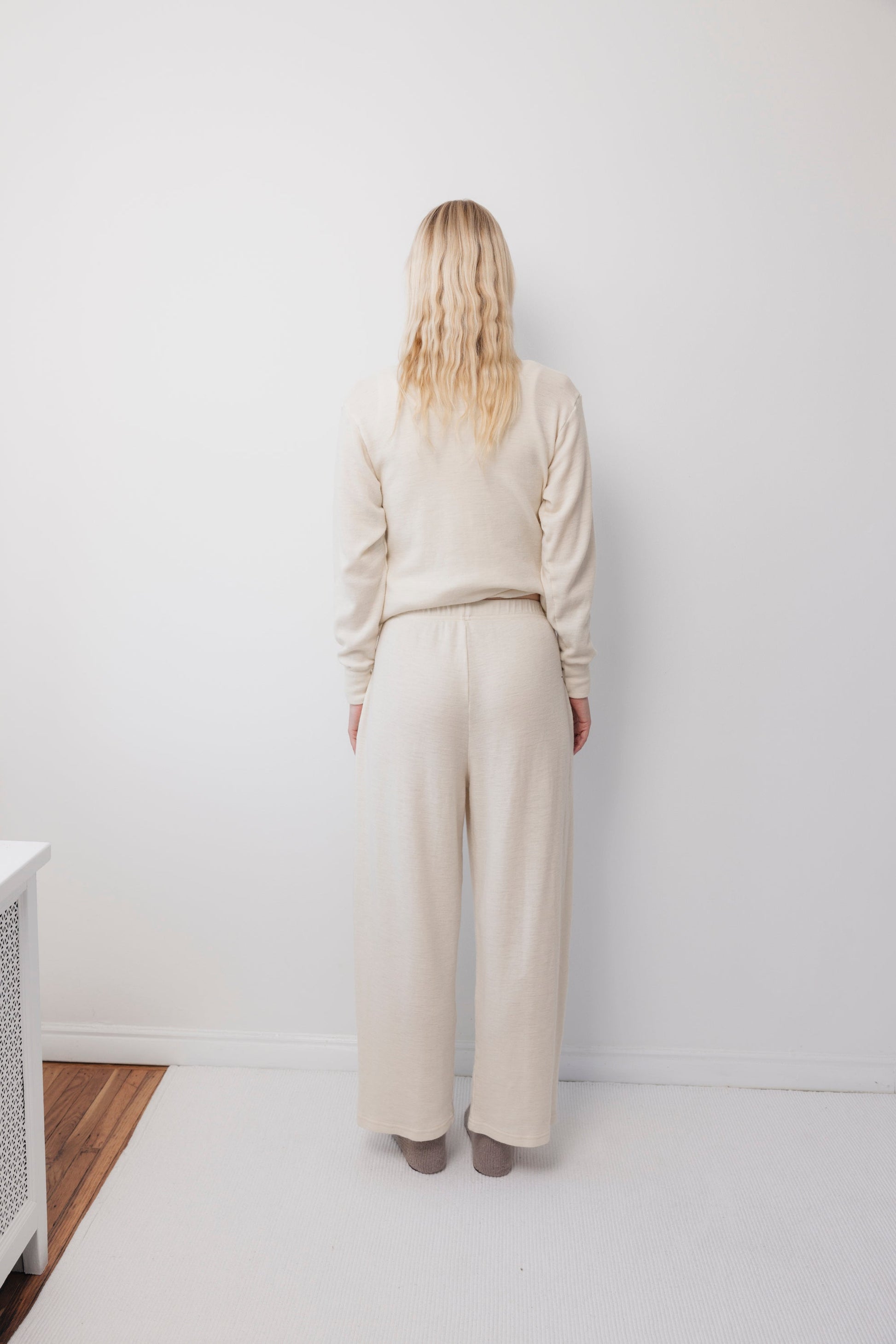 A person with long blonde hair stands facing a white wall, wearing Wol Hide's Wool Wide Leg: Natural pants and a matching cream-colored long-sleeve top, on a white carpeted floor.