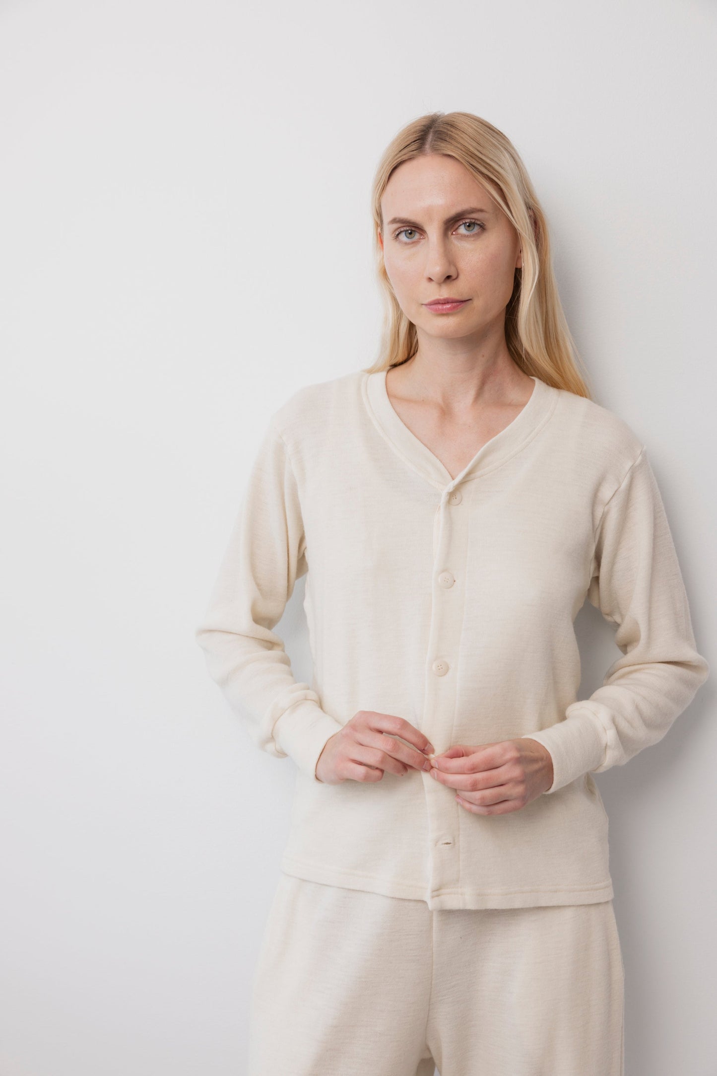 A woman with long blonde hair stands against a white background, wearing Wol Hide’s Wool Liner : Natural cardigan with corozo buttons and matching pants, looking calmly at the camera with her hands clasped.