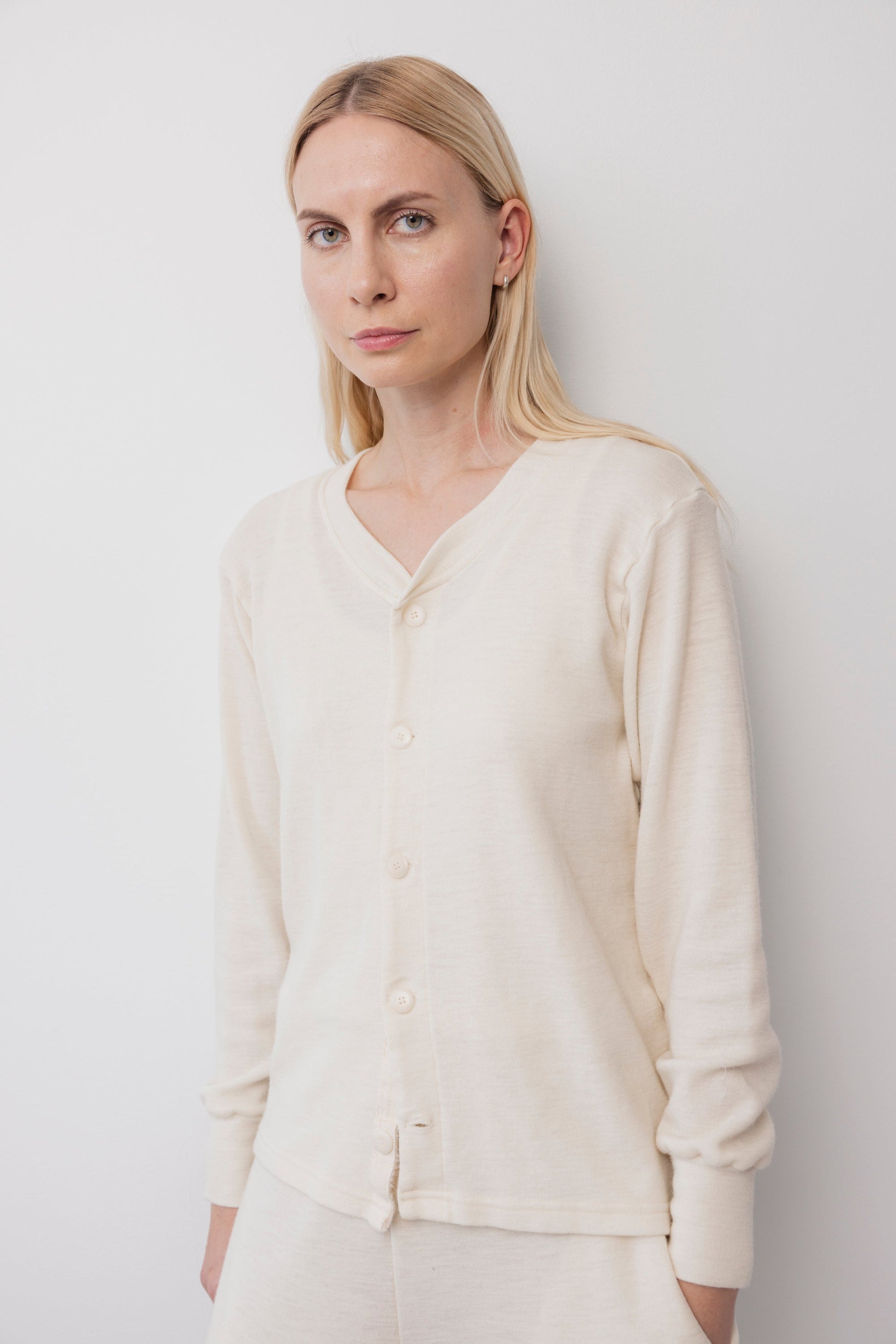 A woman with long blonde hair wears the Wol Hide Wool Liner : Natural, a cream long-sleeve slim cardigan with Corozo buttons, standing calmly against a plain white wall.