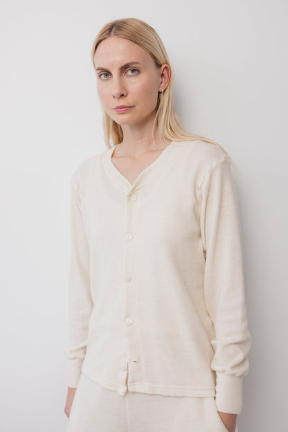A woman with long blonde hair wears the Wol Hide Wool Liner : Natural, a cream long-sleeve slim cardigan with Corozo buttons, standing calmly against a plain white wall.