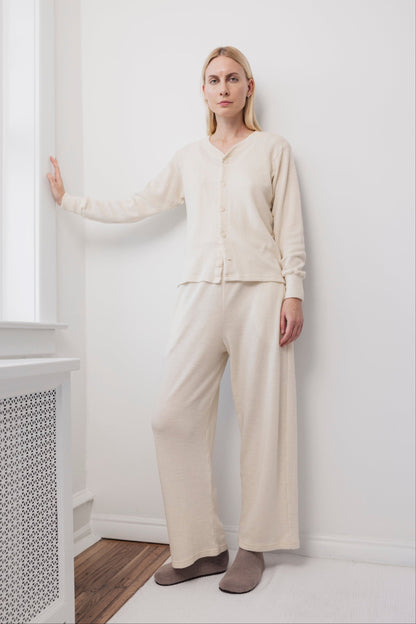 A woman stands indoors against a white wall, wearing the Wol Hide Wool Wide Leg pants in Natural, paired with a cream-colored cardigan and beige slippers. She has blonde hair and a calm expression, one hand resting on the wall beside a window.