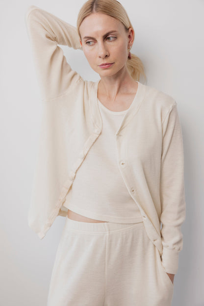 A blonde woman wears the Wol Hide Wool Liner cardigan in Natural with matching pants, featuring lightweight wool and Corozo buttons. She stands against a white background, one arm raised to her head, gazing to the side.