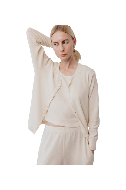 A woman in cream Wol Hide Wool Liner loungewear stands with one hand behind her head, looking to the side. She wears a matching set that embodies minimalist style against a plain white background.