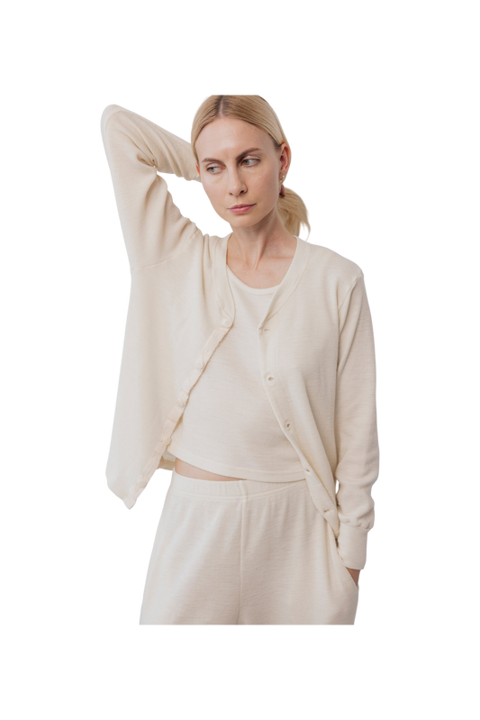A woman in cream Wol Hide Wool Liner loungewear stands with one hand behind her head, looking to the side. She wears a matching set that embodies minimalist style against a plain white background.