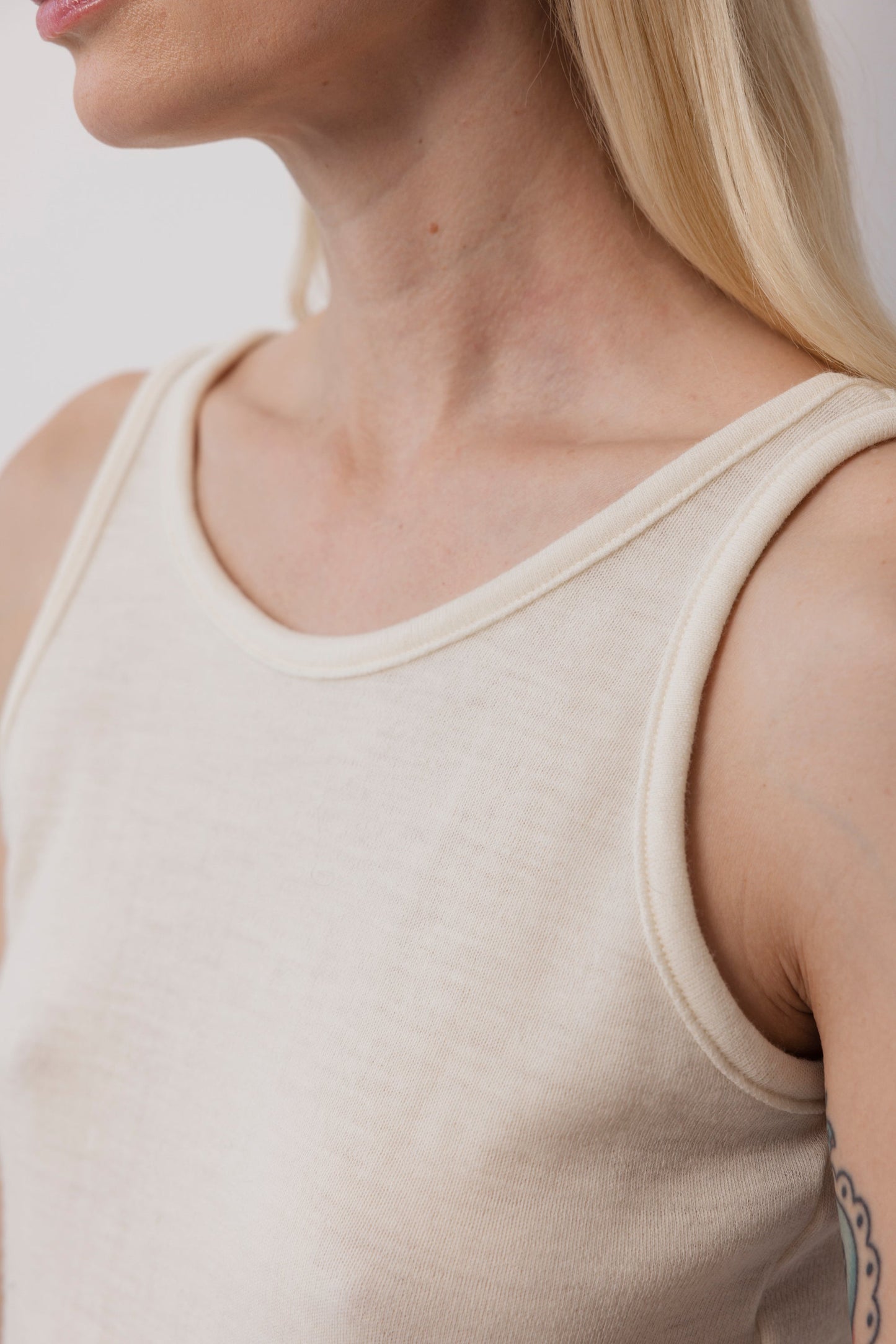 A close-up shows a person with long blonde hair wearing the Wol Hide Wool Tank: Natural. Their shoulders, upper chest, and part of an upper arm tattoo are visible against a plain light background.