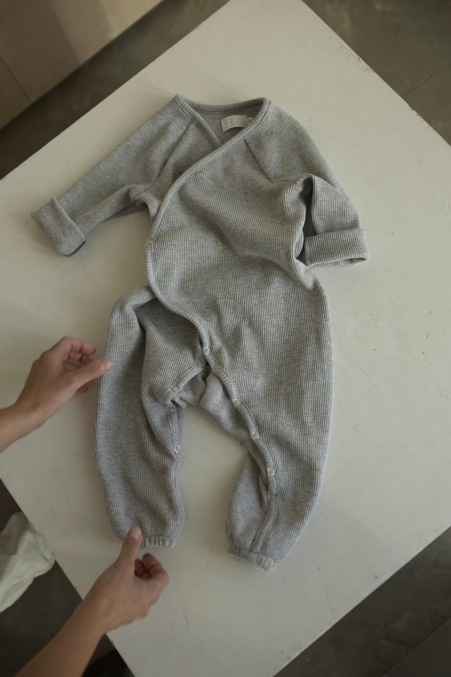 An individual arranges the cabane childrenswear Après Wrap Organic Waffle, an ethically handmade gray baby onesie with long sleeves and buttons, on a white surface. The well-lit setting highlights its soft, textured fabric and intricate details.
