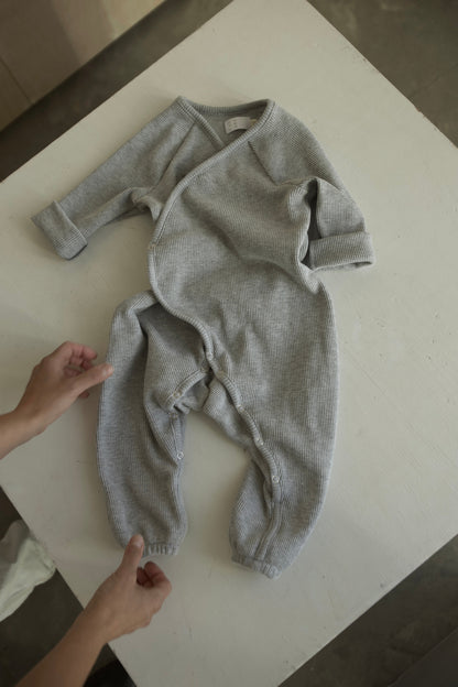 An individual arranges the cabane childrenswear Après Wrap Organic Waffle, an ethically handmade gray baby onesie with long sleeves and buttons, on a white surface. The well-lit setting highlights its soft, textured fabric and intricate details.
