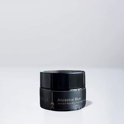 A small black jar labeled “Ancestral Blue 2.4oz” by Wild Mamas rests on a white surface with a light gray background. The label highlights Manuka, Royal Jelly, Blue Tansy, and grass-fed tallow for natural skincare. Net weight: 2.4 oz.