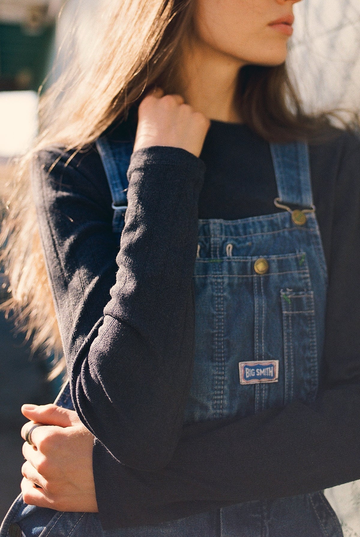 A person with long brown hair wears a black shirt under denim overalls from "Big Smith," with one arm crossed and the other near their collar, revealing a hint of LA RELAXED's "Organic Texture Long Sleeve." Sunlight casts soft shadows on the label.
