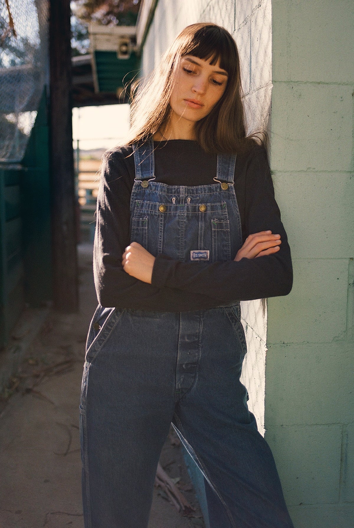 A person with long brown hair stands against a light green wall wearing blue denim overalls and an LA RELAXED Organic Texture Long Sleeve shirt in black. They cross their arms thoughtfully as sunlight casts shadows on the concrete ground.