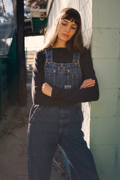 A person with long brown hair stands against a light green wall wearing blue denim overalls and an LA RELAXED Organic Texture Long Sleeve shirt in black. They cross their arms thoughtfully as sunlight casts shadows on the concrete ground.