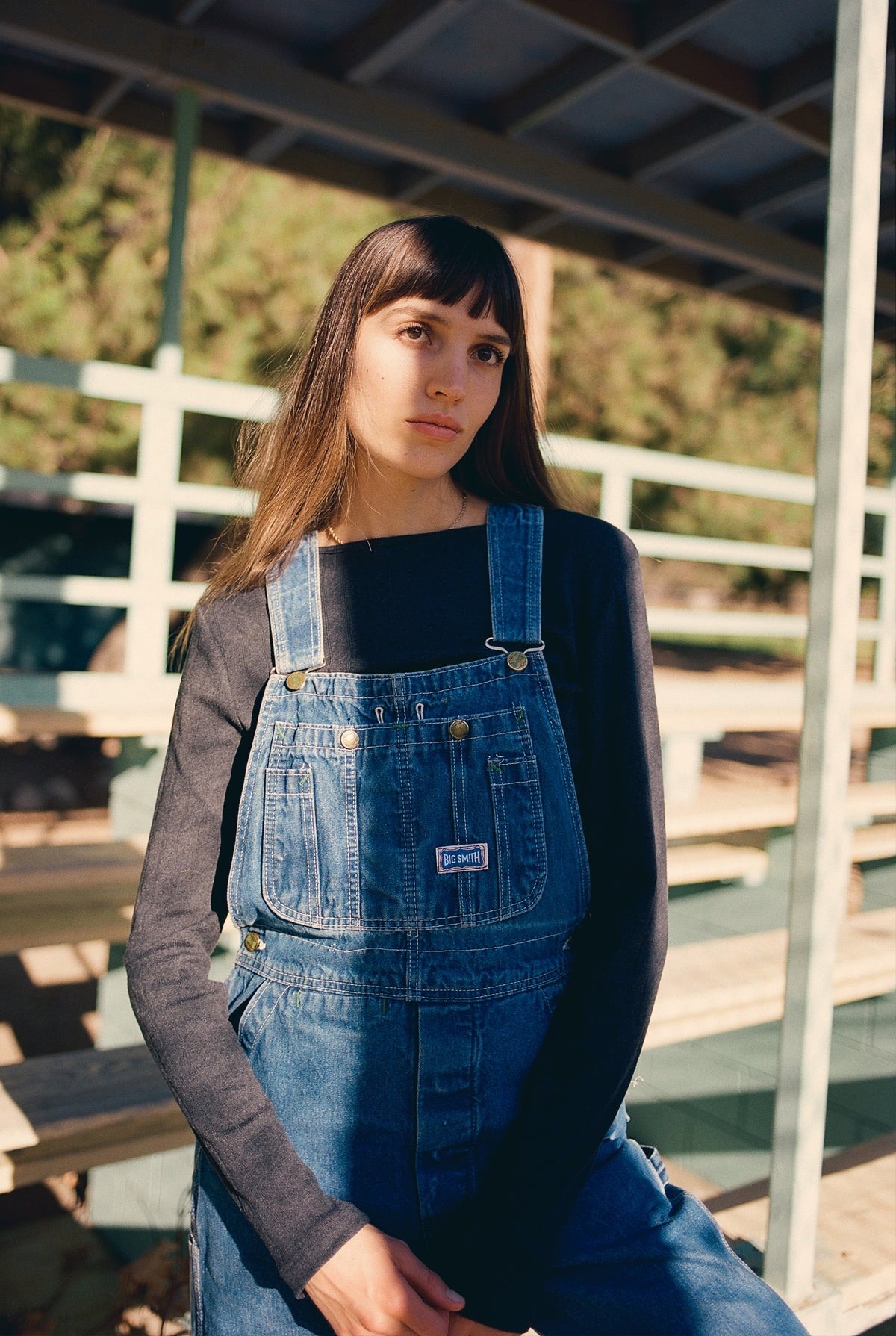 A person with long hair wears an LA RELAXED Organic Texture Long Sleeve under denim overalls, standing outside beneath a structure with green railings and seating. The serene backdrop features trees and soft natural lighting.