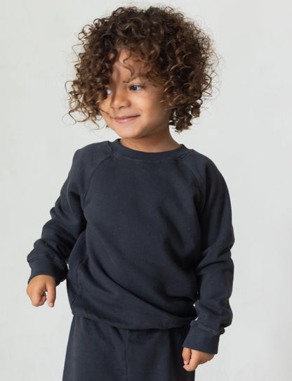 A young child with curly hair smiles and looks off to the side, wearing The Lil' Classic Crewneck and matching pants made of organic French terry cloth by all the babies, against a plain white background.