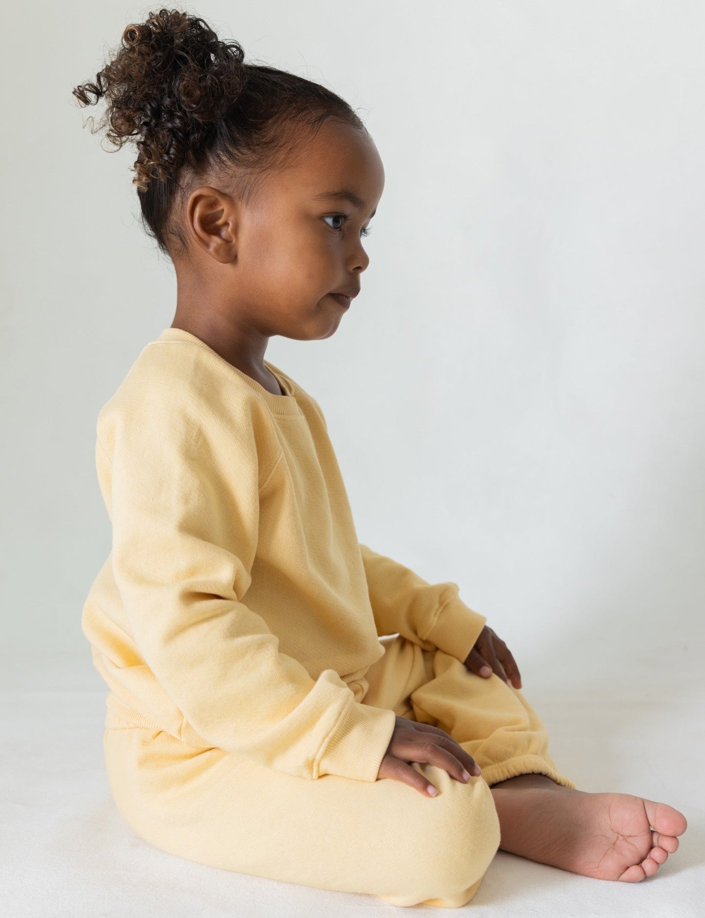 Butter -
A young child with curly hair sits cross-legged on the floor, wearing The Lil' Classic Sweatpant from all the babies, crafted from organic French terry cloth. The matching light yellow sweatshirt and vintage-inspired sweatpants enhance their serene expression against the plain, light-colored background that focuses attention on their side profile.