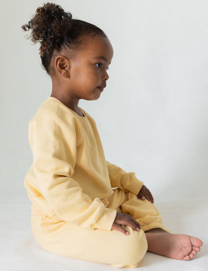 Butter -
A young child with curly hair sits cross-legged on the floor, wearing The Lil' Classic Sweatpant from all the babies, crafted from organic French terry cloth. The matching light yellow sweatshirt and vintage-inspired sweatpants enhance their serene expression against the plain, light-colored background that focuses attention on their side profile.
