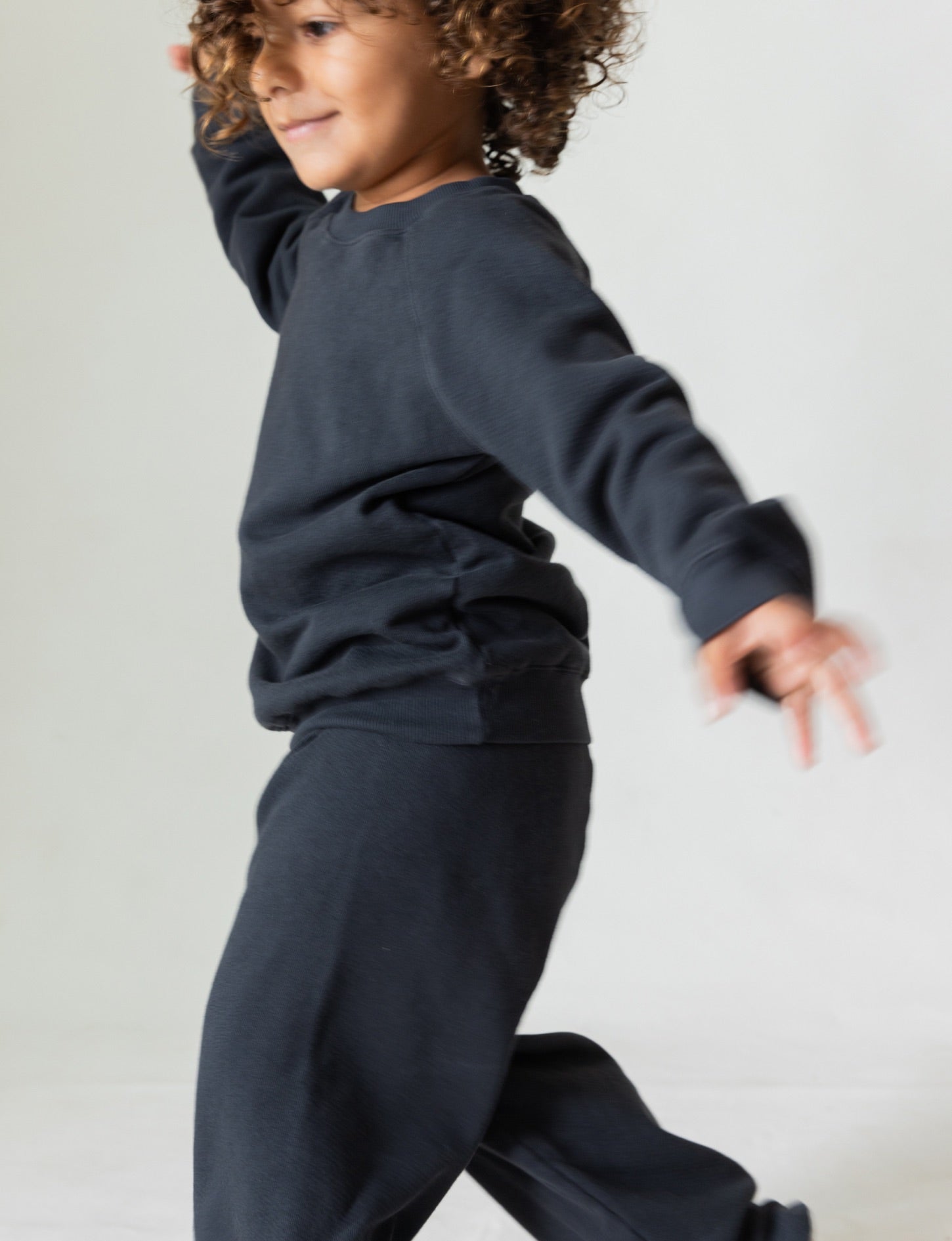 A young child with curly hair is smiling and dancing in a matching dark outfit made from organic French terry cloth, consisting of The Lil' Classic Crewneck top and pants from all the babies. The background is plain and light-colored.