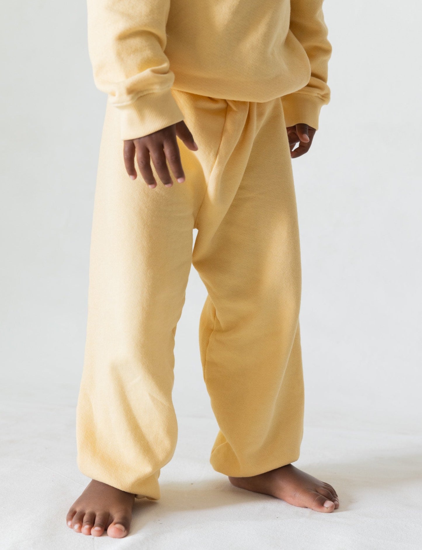 Butter - 
A young child stands barefoot on a white surface, wearing an oversized pale yellow sweat suit made from organic French terry cloth. The outfit includes a sweatshirt and The Lil' Classic Sweatpant by all the babies, slightly too big for the child, creating a cozy and relaxed appearance. The child's hands and feet are visible.