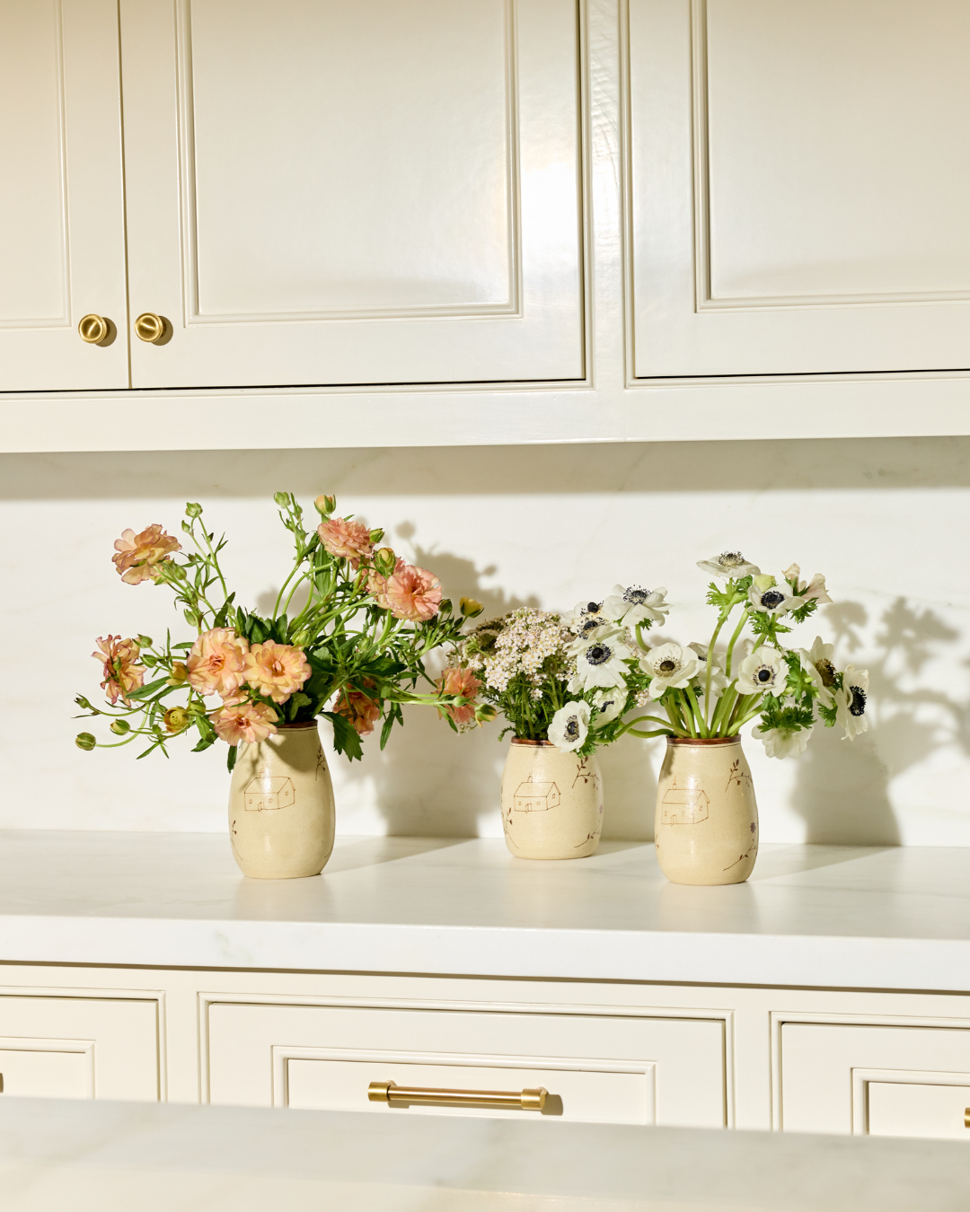 Three cream-colored Baile Homestead Vases by Baile, featuring hand-drawn designs, are filled with pink and white flowers and displayed on a white kitchen counter before cream cabinetry with gold handles.