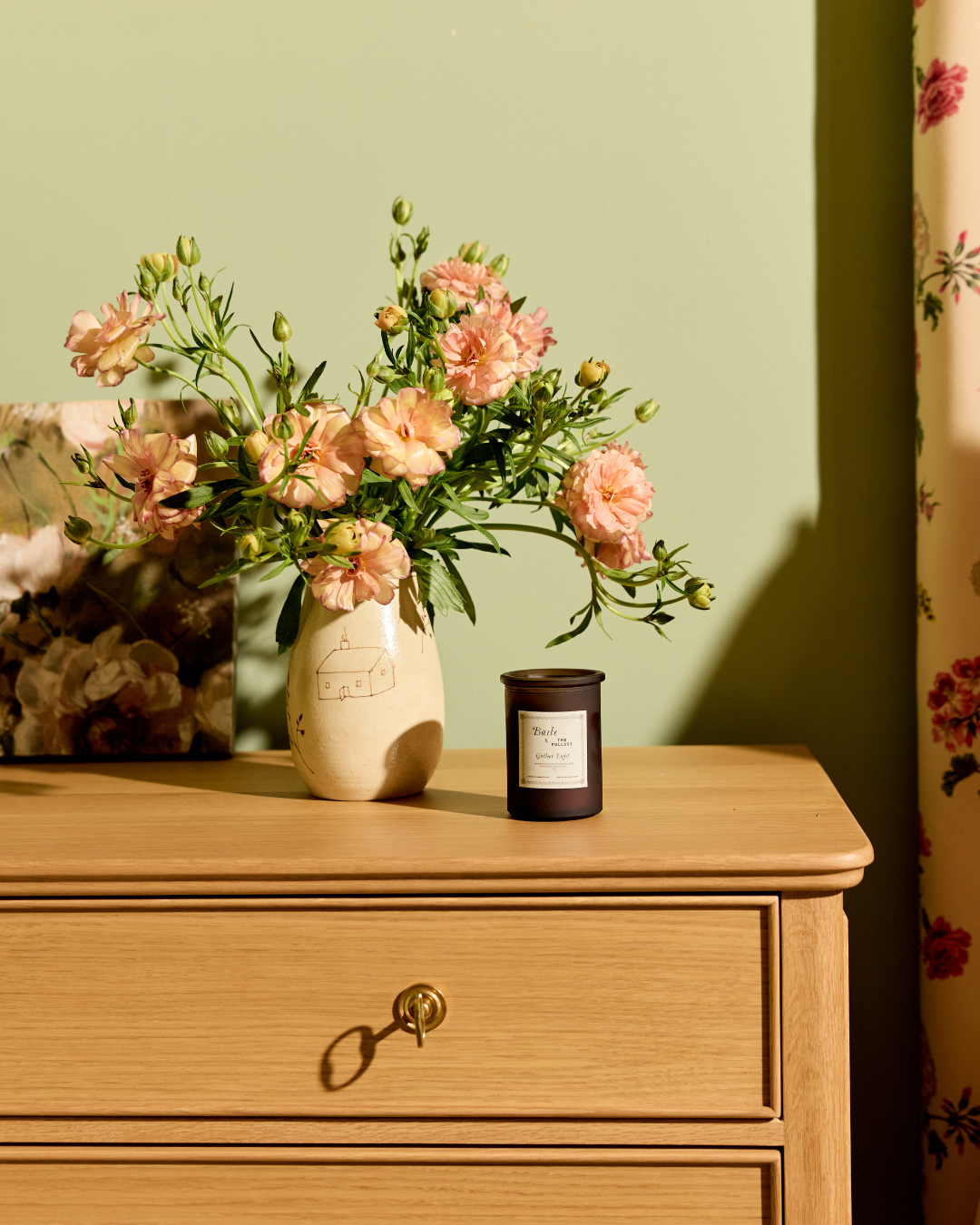 A wooden dresser with a key in the drawer features the Baile Homestead Vase by Baile, filled with pink flowers, and a dark candle on top. The setup sits against a green wall with floral curtains lit by gentle sunlight.