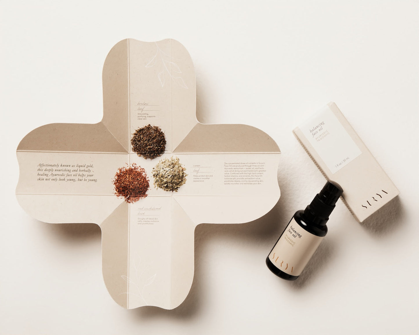 A flat-lay photograph showcasing a skincare package from Surya. The contents include a black spray bottle labeled "Balancing Face Oil," an open box with text and graphics, and small piles of dried herbs in different colors. The minimalist beige and white designs on the box and bottle exude elegance.