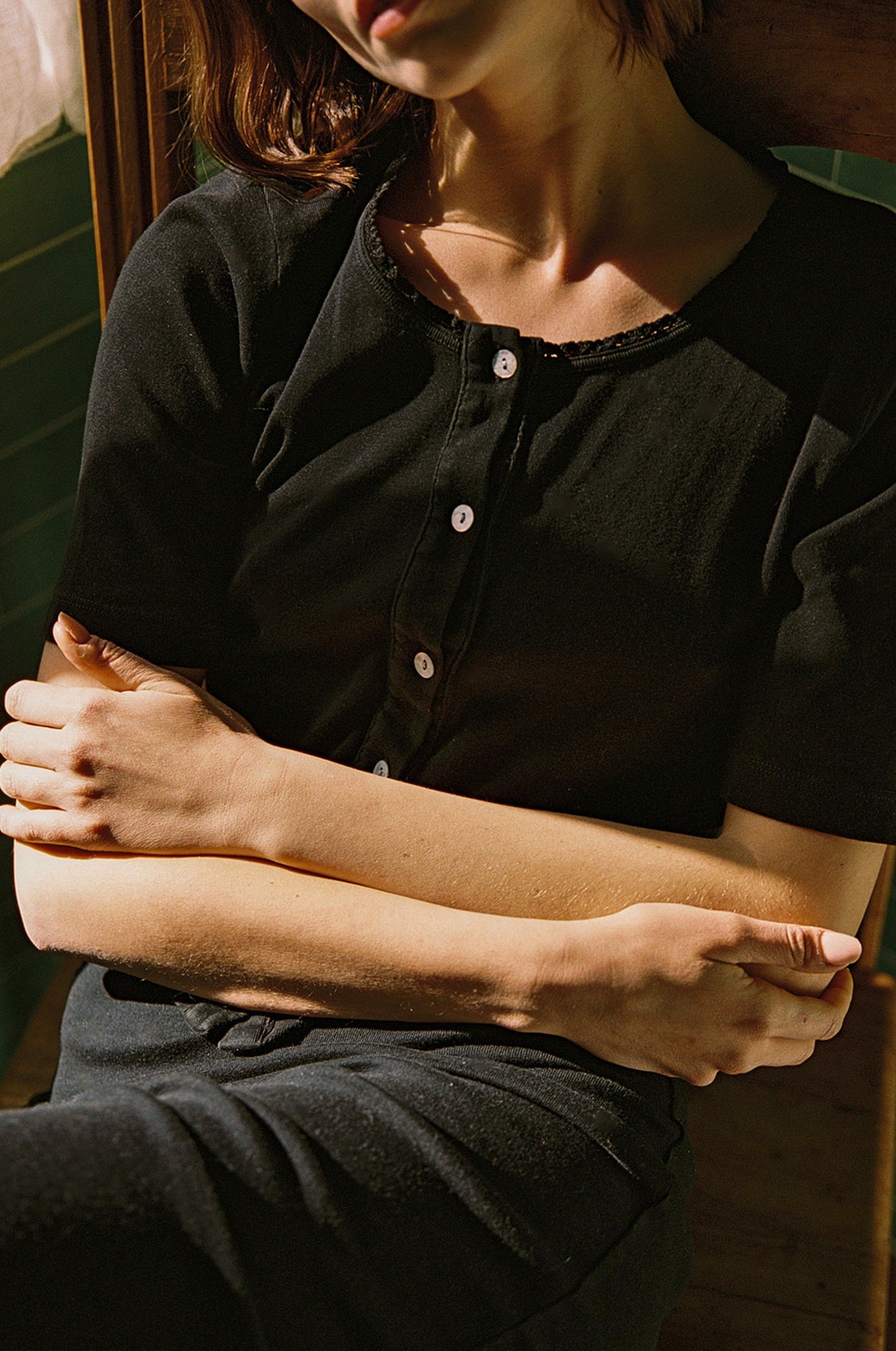 A person wearing an LA Relaxed Short Sleeve Organic Cardi sits with arms crossed in a sunlit area that casts intricate shadows on their face.