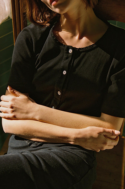 A person wearing an LA Relaxed Short Sleeve Organic Cardi sits with arms crossed in a sunlit area that casts intricate shadows on their face.
