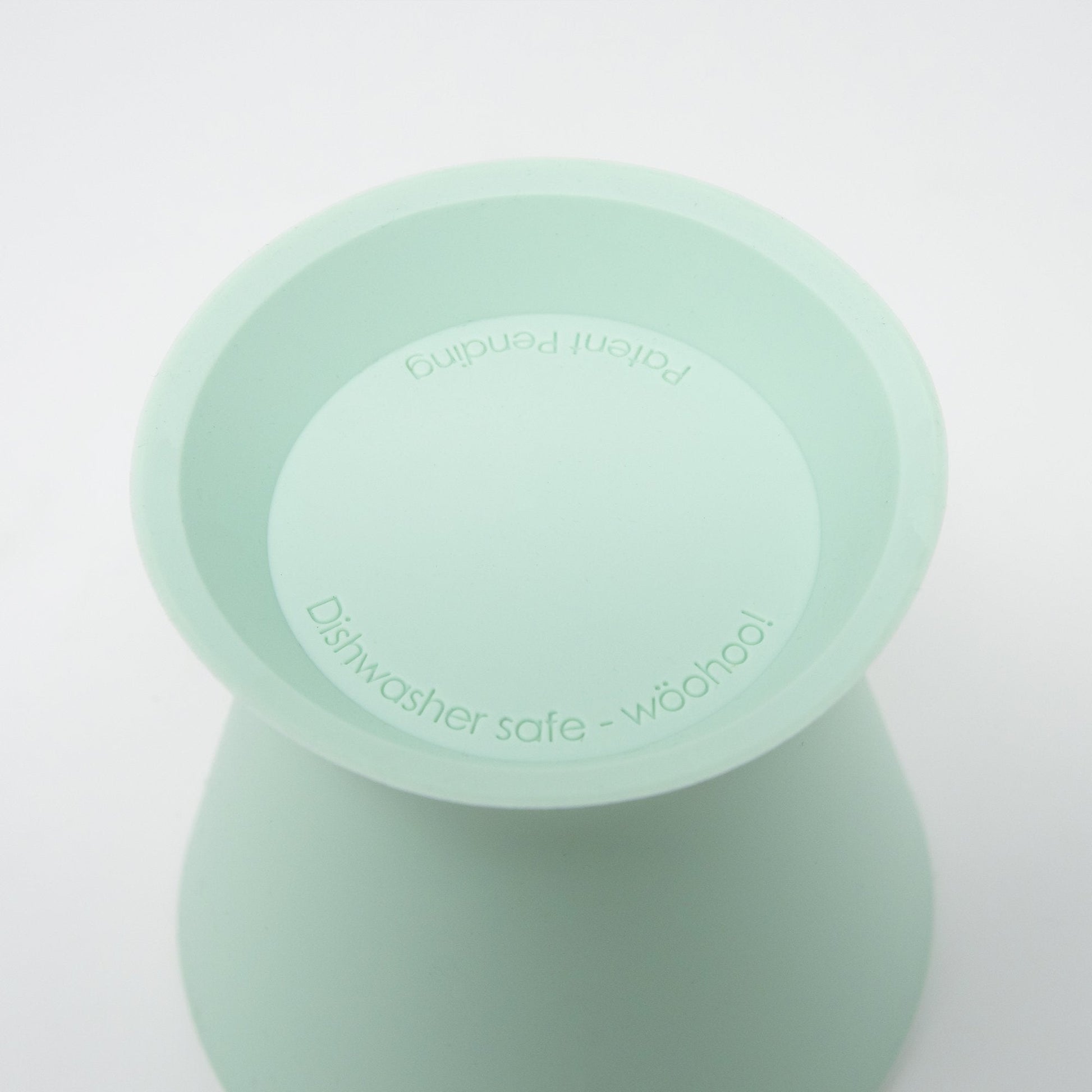 Close-up of a light green BPA-free silicone bökee Baby Bottle Holder by bökee, featuring curved sides and a flat top. The text inside reads "Dishwasher safe - wööhoo!" and "Patent pending." Ideal for one-handed bottle prep, set against a plain white background.