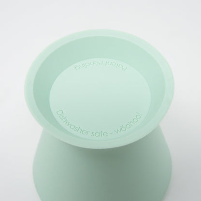 Close-up of a light green BPA-free silicone bökee Baby Bottle Holder by bökee, featuring curved sides and a flat top. The text inside reads "Dishwasher safe - wööhoo!" and "Patent pending." Ideal for one-handed bottle prep, set against a plain white background.