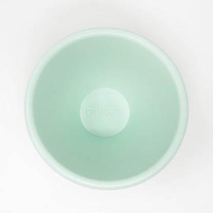 A top-down view of an empty, light green bökee Baby Bottle Holder, crafted from BPA-free silicone, shown on a white background.