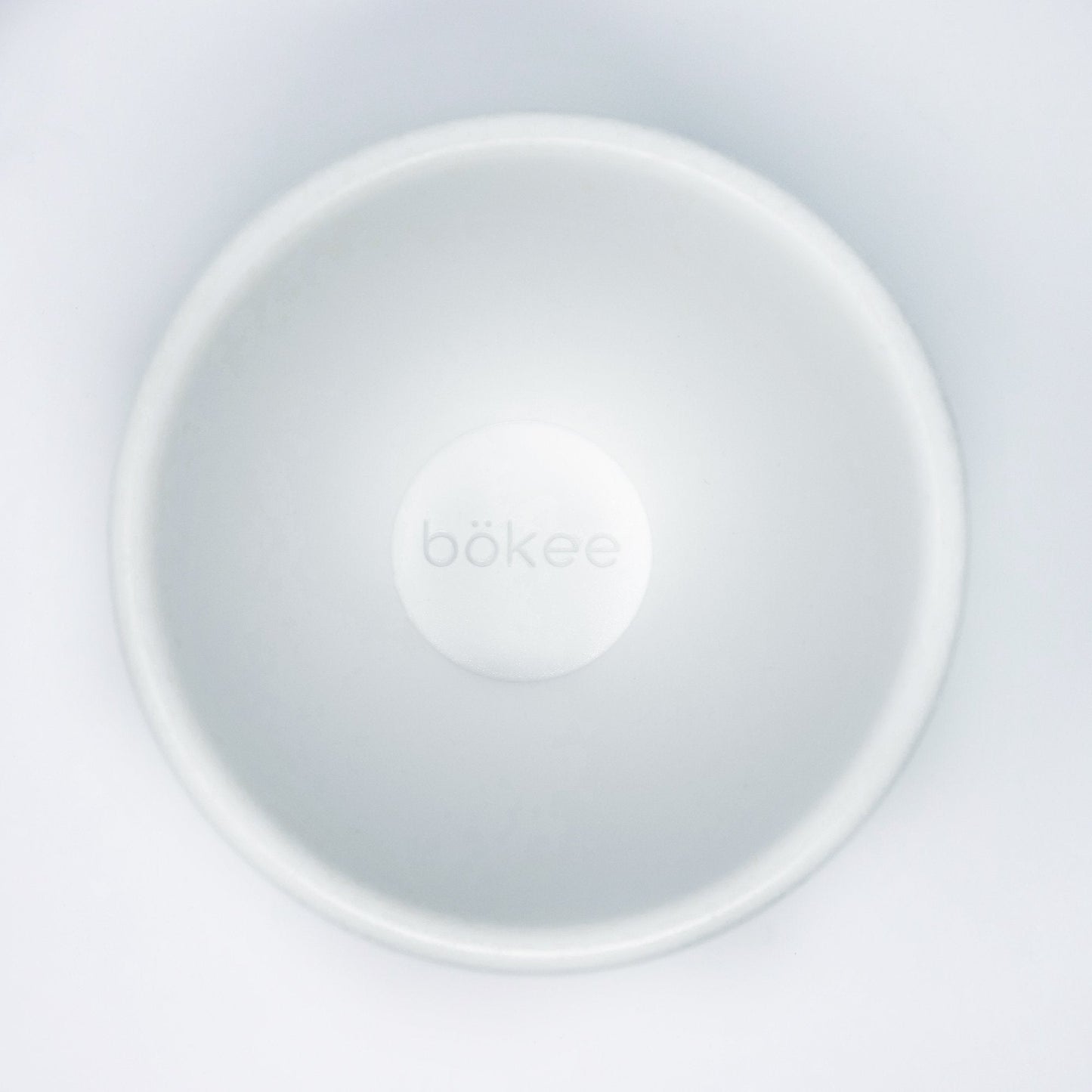 Top view of the "bökee Baby Bottle Holder, One-Handed Bottle Prep," a white, circular product crafted from BPA-free silicone designed to hold and stabilize baby bottles during one-handed bottle preparation. The smooth, minimalist design prominently features the brand name "bökee" in the center.