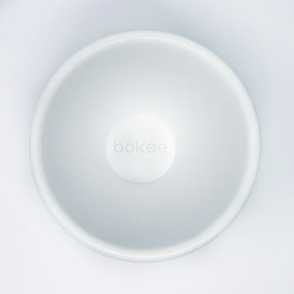 Top view of the "bökee Baby Bottle Holder, One-Handed Bottle Prep," a white, circular product crafted from BPA-free silicone designed to hold and stabilize baby bottles during one-handed bottle preparation. The smooth, minimalist design prominently features the brand name "bökee" in the center.