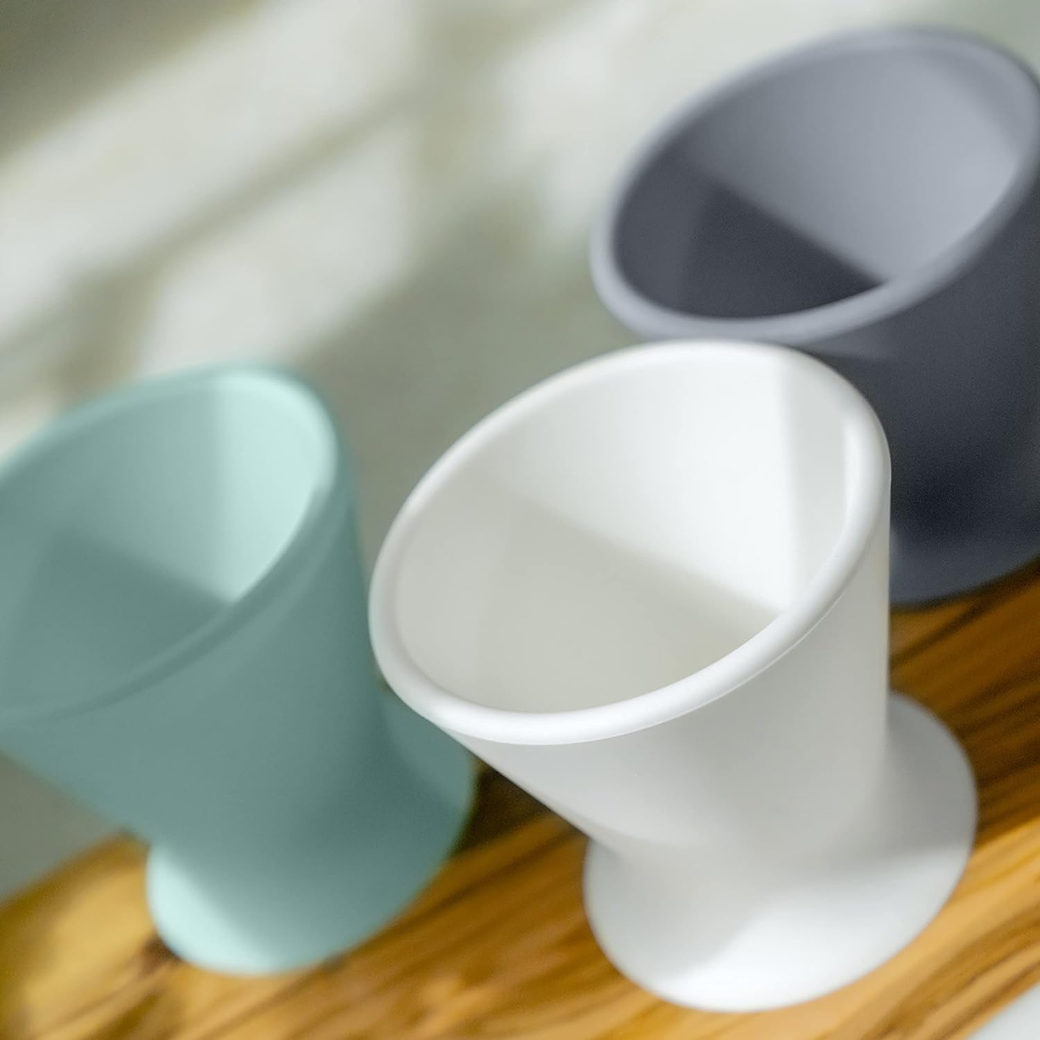 Three minimalist, matte-finished vases in pastel colors (mint green, white, and gray) are arranged on a wooden surface. Resembling the sleek design of the bökee Baby Bottle Holder by bökee, these vases boast gentle curves. Soft lighting casts subtle shadows in the background.