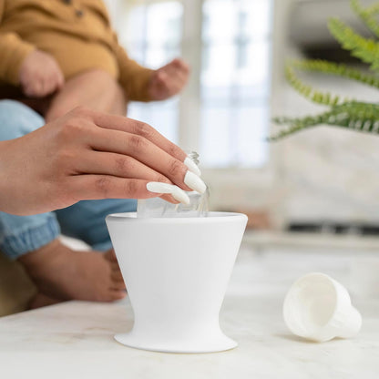 A person with long, white nails skillfully pours liquid from a BPA-free silicone bottle into a sleek, white humidifier. Nearby, a child sits close to the bökee Baby Bottle Holder by bökee, while a plant and baby bottles are subtly visible on the marble surface in the background.