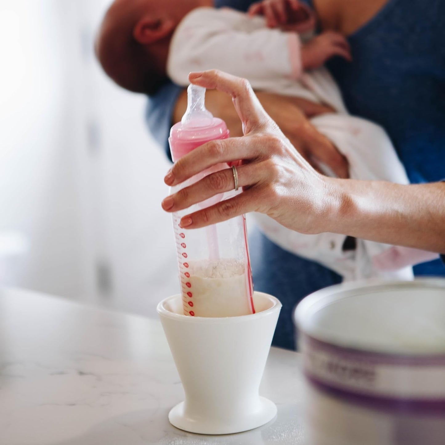 A person is expertly using the bökee Baby Bottle Holder for a one-handed bottle prep with BPA-free silicone baby bottles. As they hold a baby dressed in light-colored clothing, they fill the bottle with formula powder from the nearby container, ready for use.